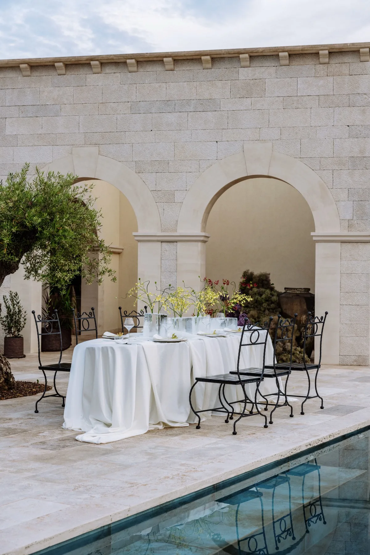 A white draped table set beneath the stone arches beside the pool at LaMaison